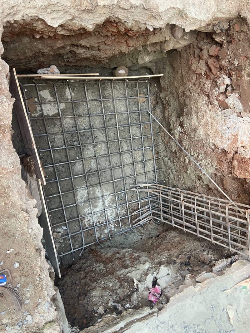 Top-down view into a footing pit showing a square grid of reinforcement bars and a column cage lying alongside.
