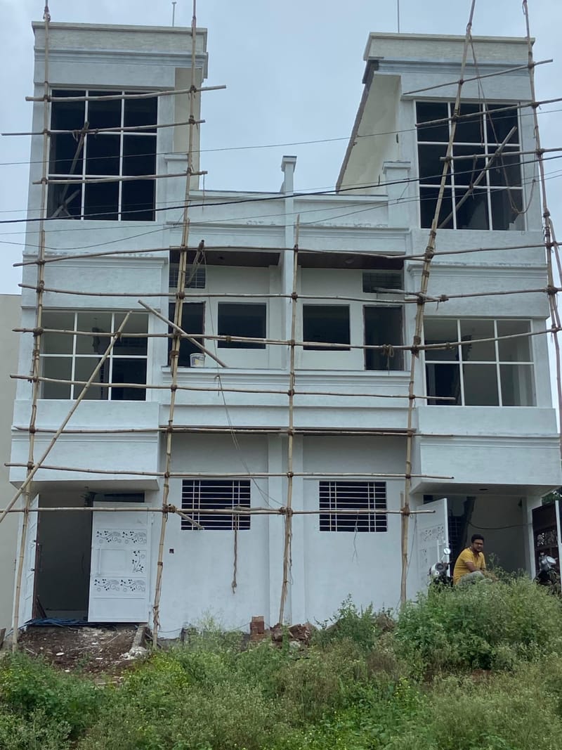 The same house on site, painted white with gridded windows installed in both corner bays and bamboo scaffolding across the front.